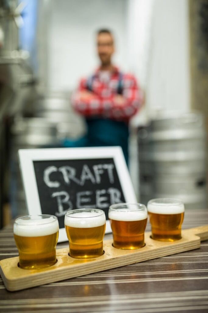 Sampler tray of four craft beers with varying colours and foam, foregrounded in a Barossa Valley microbrewery with a brewer standing in the background.