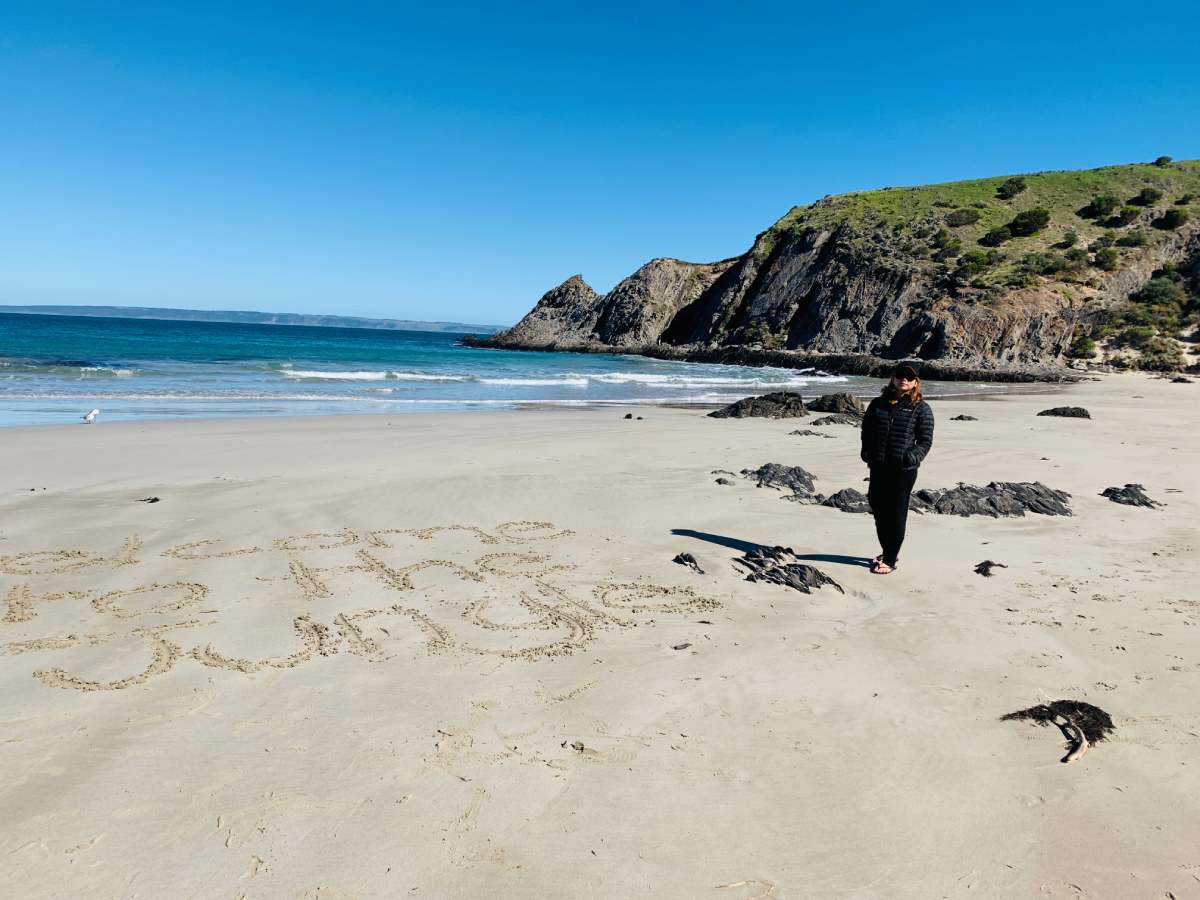 blowhole beach (1) A woman walking along the sandy shore of Blowhole Beach, surrounded by rugged cliffs and clear blue water, showcasing the natural beauty of one of the best beaches in Fleurieu Peninsula.