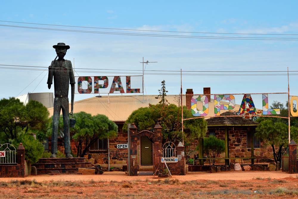 A giant miner statue standing outside an opal-themed shop in Coober Pedy, with colourful signage and rustic architecture. This iconic location highlights Coober Pedy's unique charm as one of the best historical sites in the outback, known for its opal mining heritage and underground living.