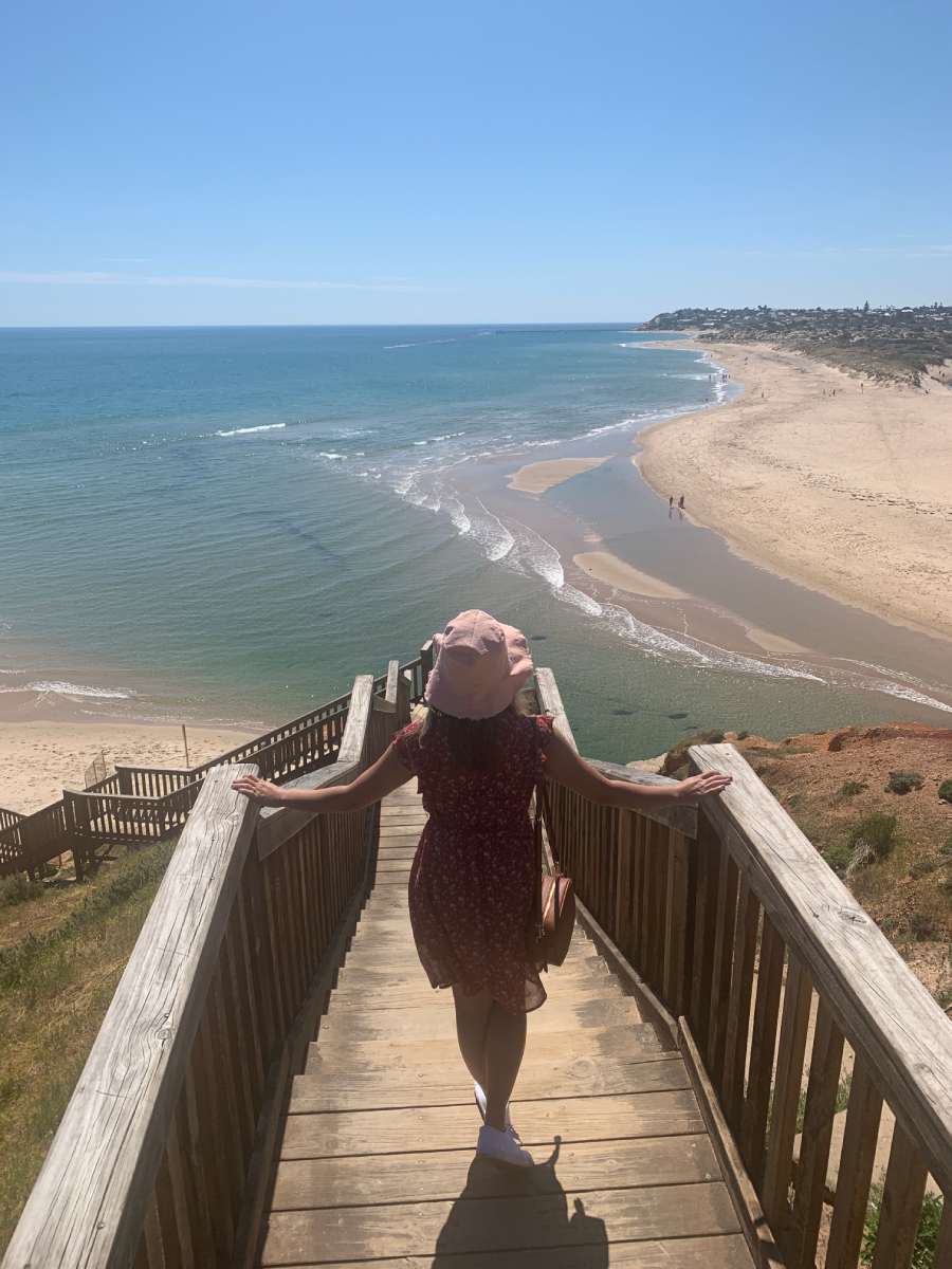 southport beach A woman walking down a wooden staircase leading to the golden sands and turquoise waters of Southport Beach, one of the must-visit beaches in the Fleurieu Peninsula.