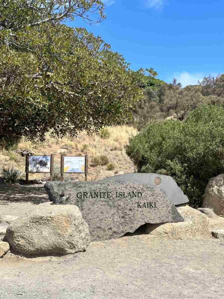 Granite Island entrance sign surrounded by native bush and information boards on a sunny day in Victor Harbor, South Australia. This scenic spot is a popular day trip from Adelaide and features among the top things to do in Adelaide in winter, especially for coastal walks, whale watching, and penguin spotting.