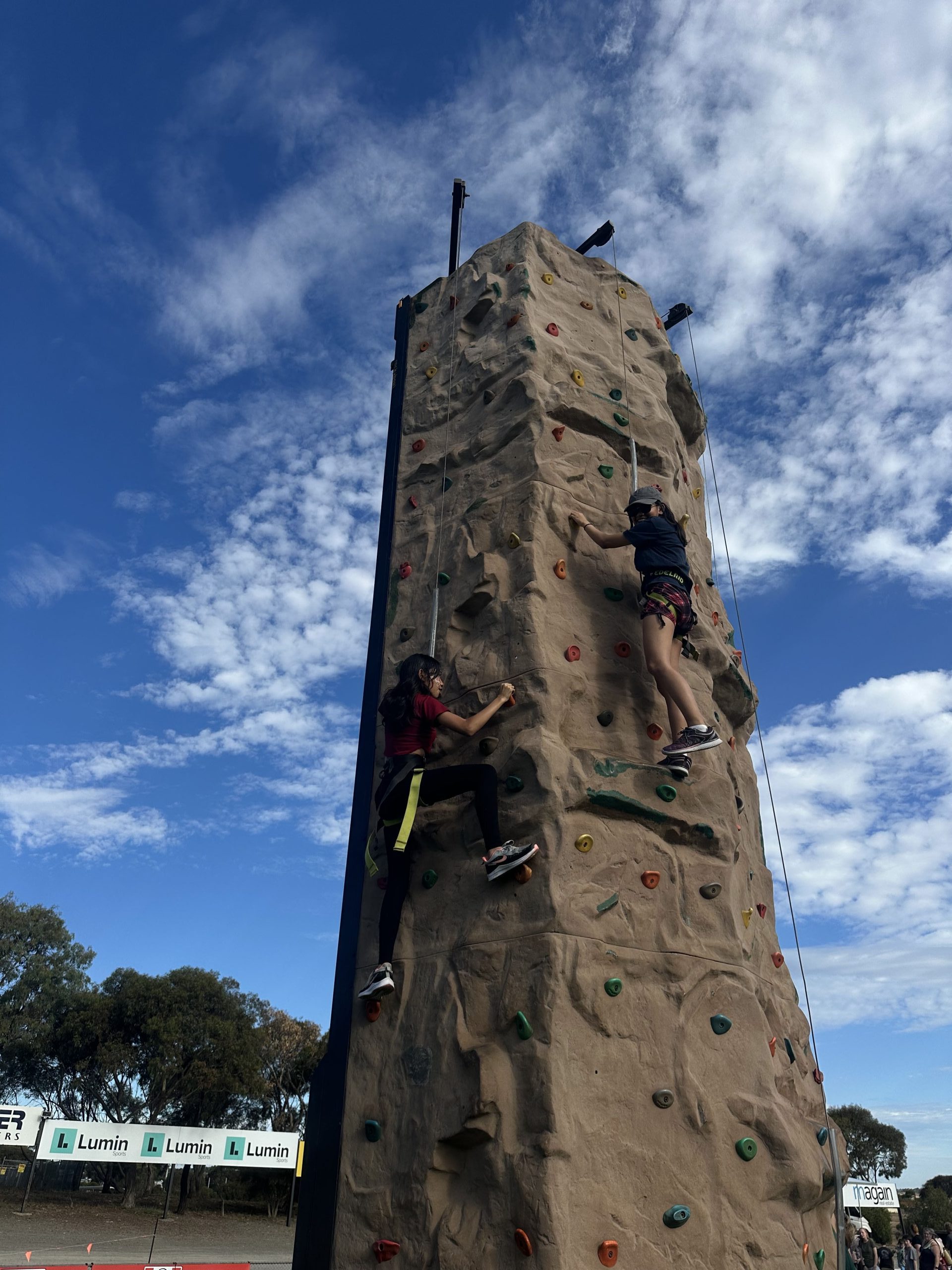 Two children climb a tall, tan-colored rock wall under a bright blue sky at an outdoor Australia Day event in Adelaide. Safety harnesses secure both climbers as they scale the wall, with colorful handholds scattered across the surface and spectators watching nearby.