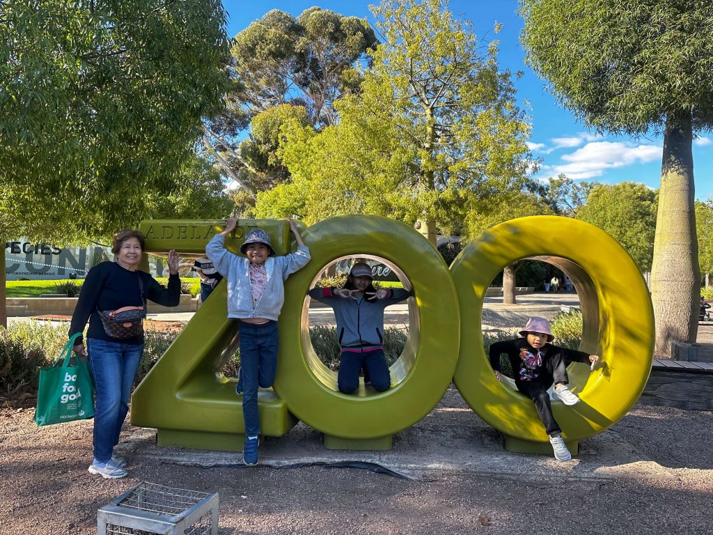 Family enjoying a day out at the Adelaide Zoo, with an older woman and three children posing and playing on the large green “ZOO” sign surrounded by trees and sunny park surroundings.