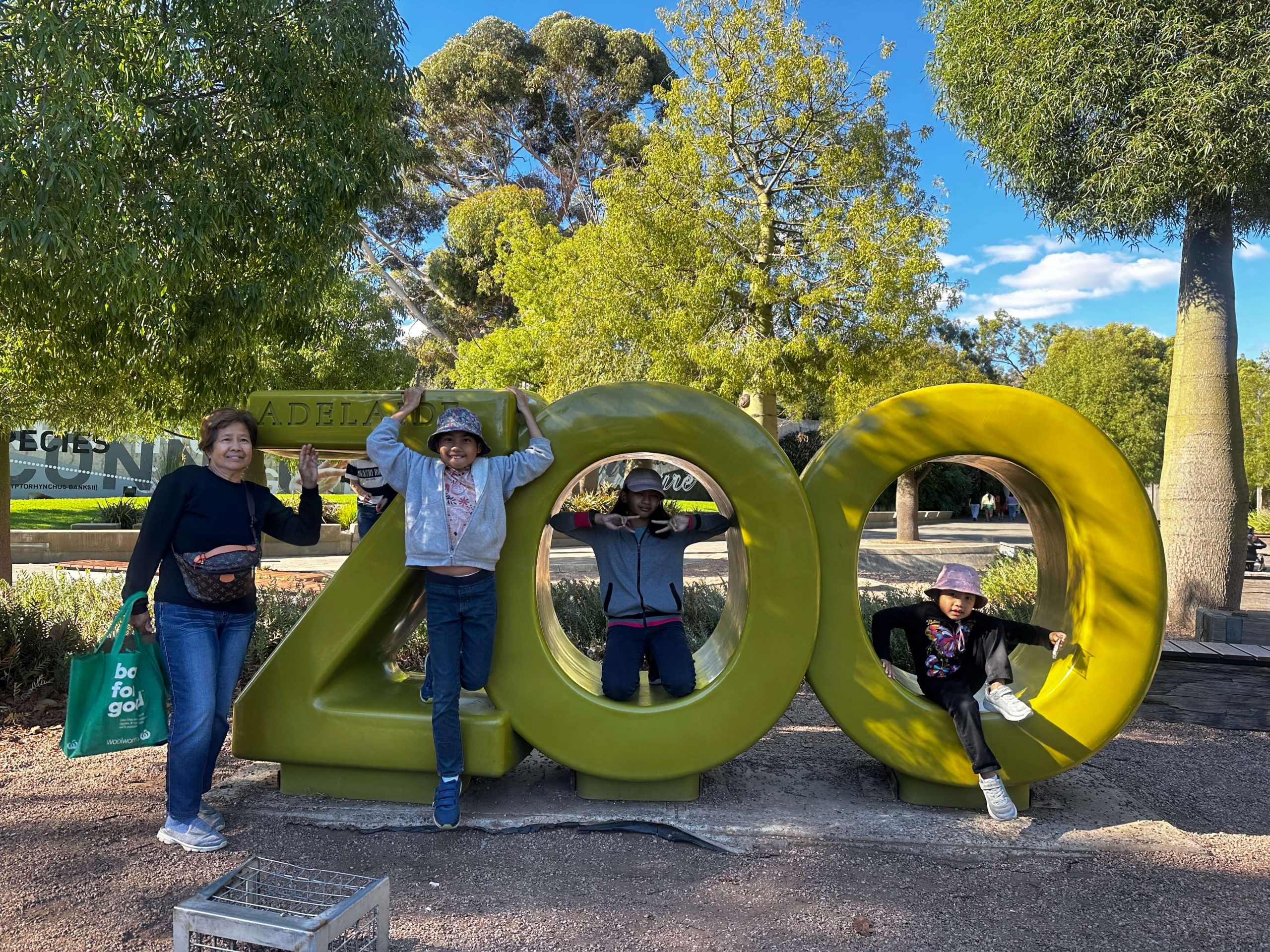 Family enjoying a day out at the Adelaide Zoo, with an older woman and three children posing and playing on the large green “ZOO” sign surrounded by trees and sunny park surroundings.