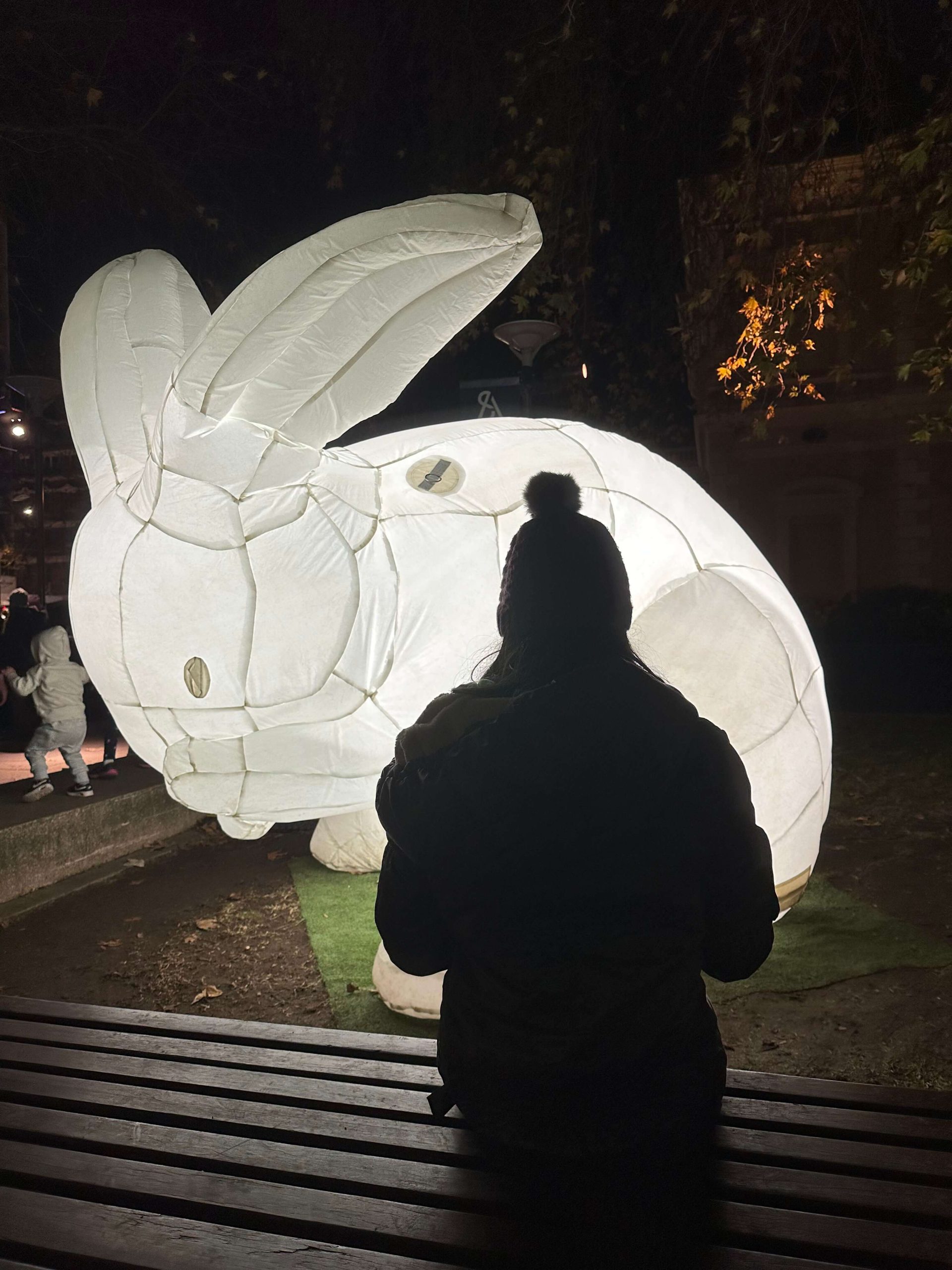 Silhouette of a person sitting on a bench in front of a giant glowing white rabbit sculpture at night during Illuminate Adelaide 2023. The illuminated rabbit lights up the dark outdoor setting while visitors walk nearby beneath hanging tree branches.