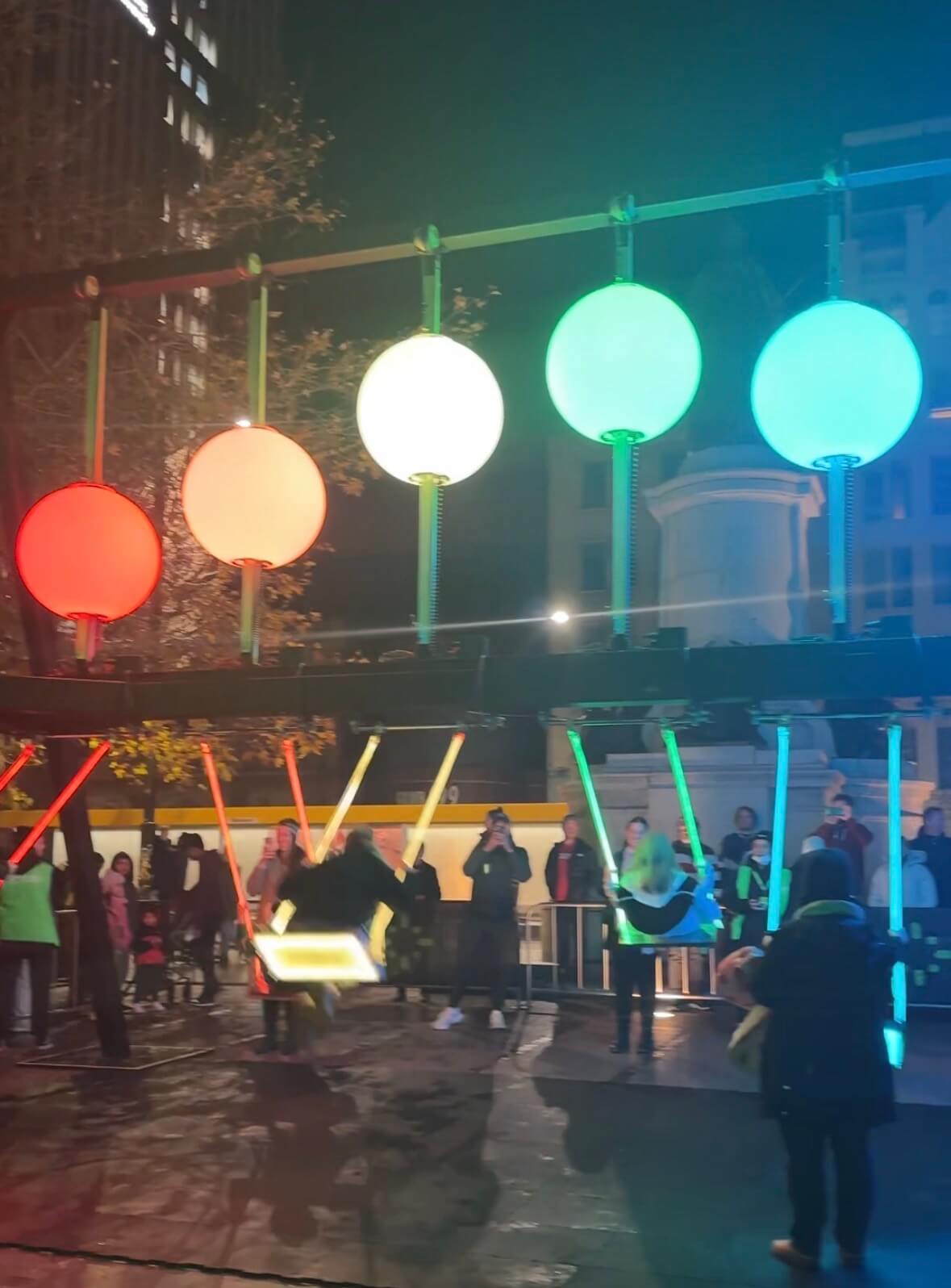 Colorful glowing swings move beneath oversized illuminated spheres in a nighttime outdoor installation. Visitors gather around the interactive display while riders swing through red, orange, white, green, and blue lights in the city square.