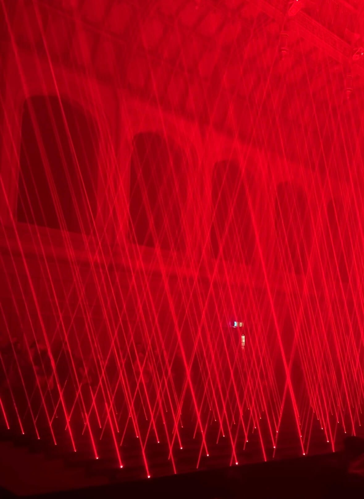 Bright red laser beams crisscross through a dark indoor space, creating a glowing geometric grid that fills the room. Silhouettes of visitors stand among the intersecting lights while haze in the air makes the laser patterns appear vivid and immersive.