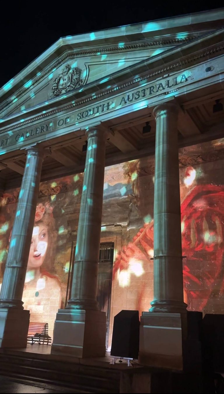 The front facade of the Art Gallery of South Australia illuminated with large-scale projected artwork and drifting light patterns at night. Tall stone columns frame the colorful projection, which features part of a painted face and rich red tones across the historic building exterior.