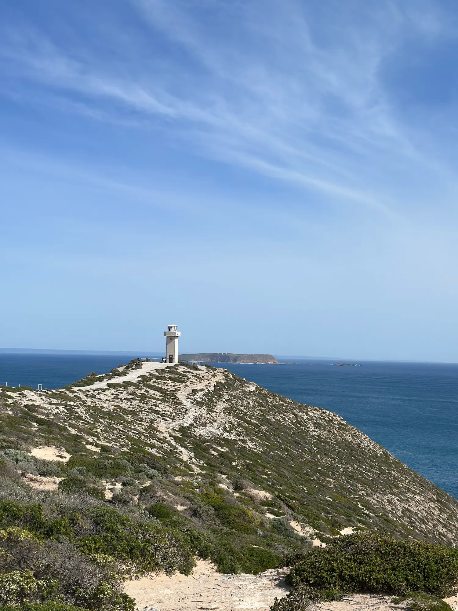 Cape Spencer Lighthouse stands on a rugged clifftop overlooking the deep blue waters of the Southern Ocean, with low coastal scrub covering the headland beneath a streaked blue sky. Cape Spencer is a spectacular stop on day trips from Adelaide, offering dramatic coastal scenery and panoramic views within Dhilba Guuranda–Innes National Park.