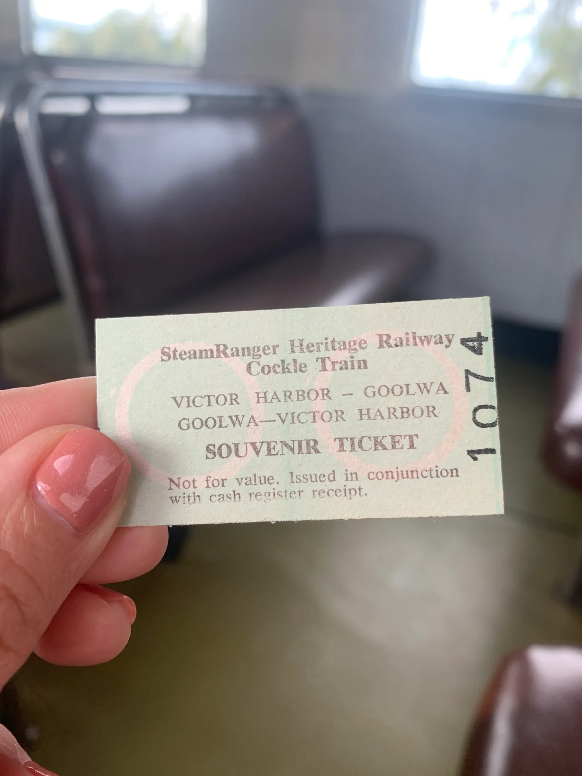 A hand holds a vintage SteamRanger Heritage Railway Cockle Train souvenir ticket inside a heritage train carriage with brown leather seats blurred in the background. The ticket reads "SteamRanger Heritage Railway Cockle Train. Victor Harbor – Goolwa. Goolwa – Victor Harbor. Souvenir Ticket. Not for value. Issued in conjunction with cash register receipt." This nostalgic rail journey is a memorable experience to include on day trips from Adelaide.