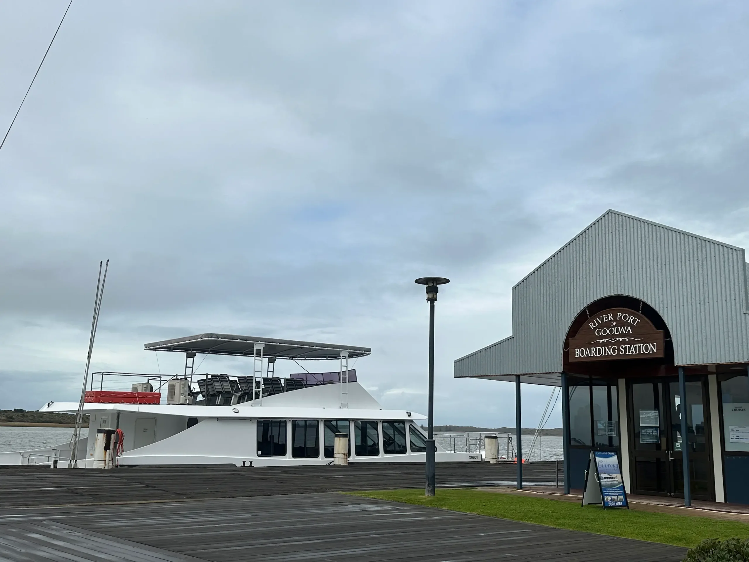 A white river cruise vessel is moored beside the River Port of Goolwa Boarding Station at Goolwa Wharf under a cloudy sky, with timber boardwalks lining the waterfront. Goolwa Wharf is a relaxing stop on day trips from Adelaide, where visitors can enjoy river cruises, historic waterfront attractions, and views of the Murray River.