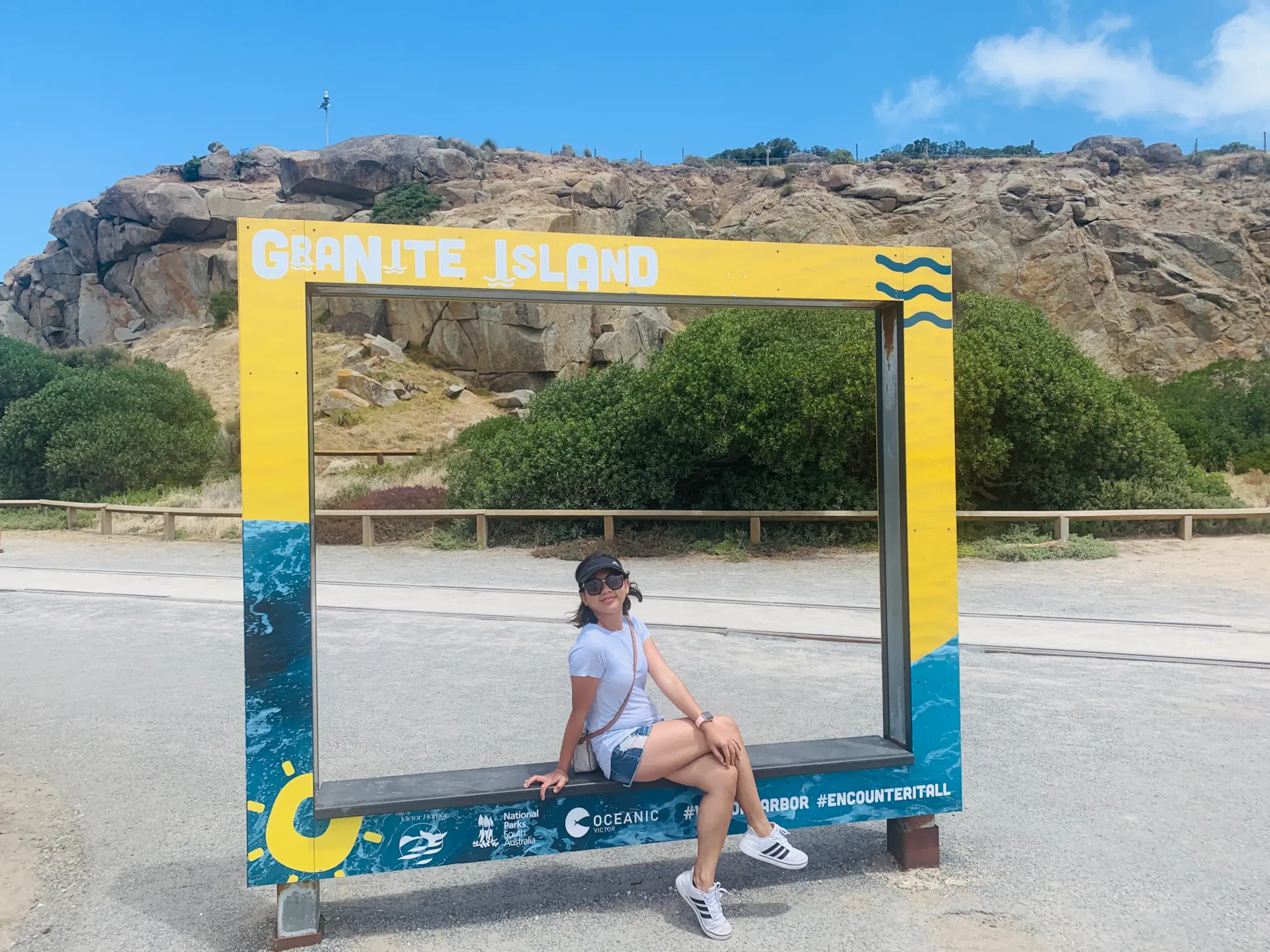 A person sits inside the bright yellow Granite Island photo frame with granite boulders and coastal scrub in the background on a sunny day. Granite Island is a scenic coastal stop for trips from Adelaide, known for its rugged shoreline, walking trails, and views across Encounter Bay.