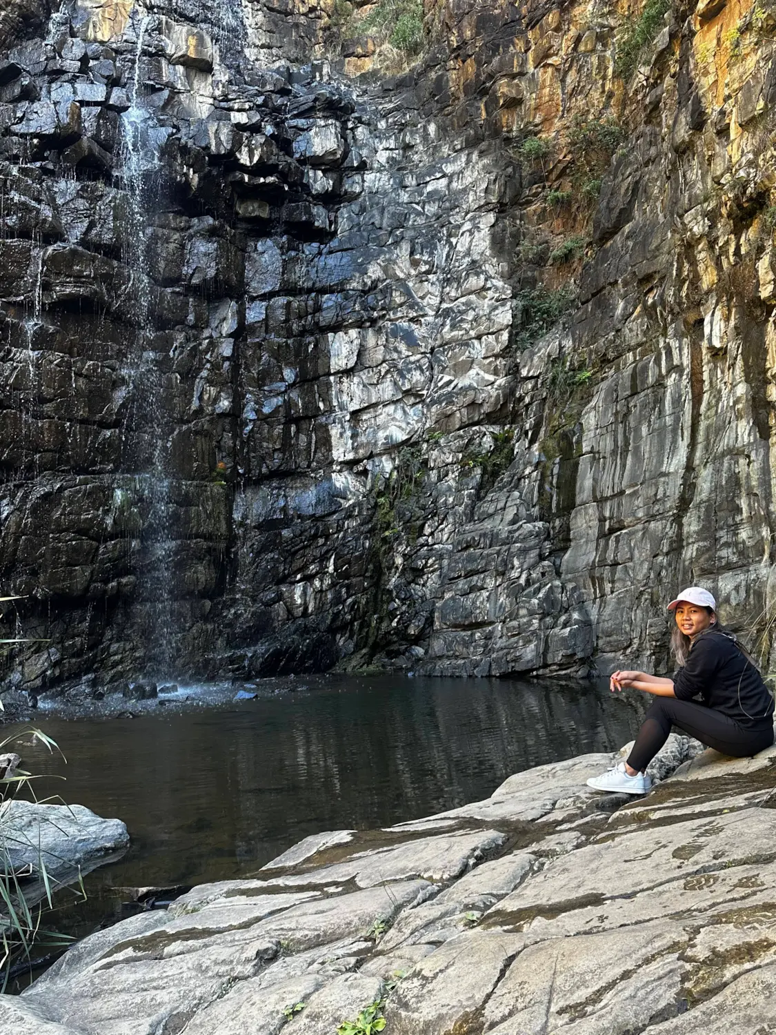 A person sits on smooth rock beside the waterfall and natural pool at Morialta Conservation Park, surrounded by towering cliff faces and rugged bushland. This peaceful hiking destination is one of the best nature escapes for day trips from Adelaide, with scenic walking trails and waterfalls after seasonal rain.