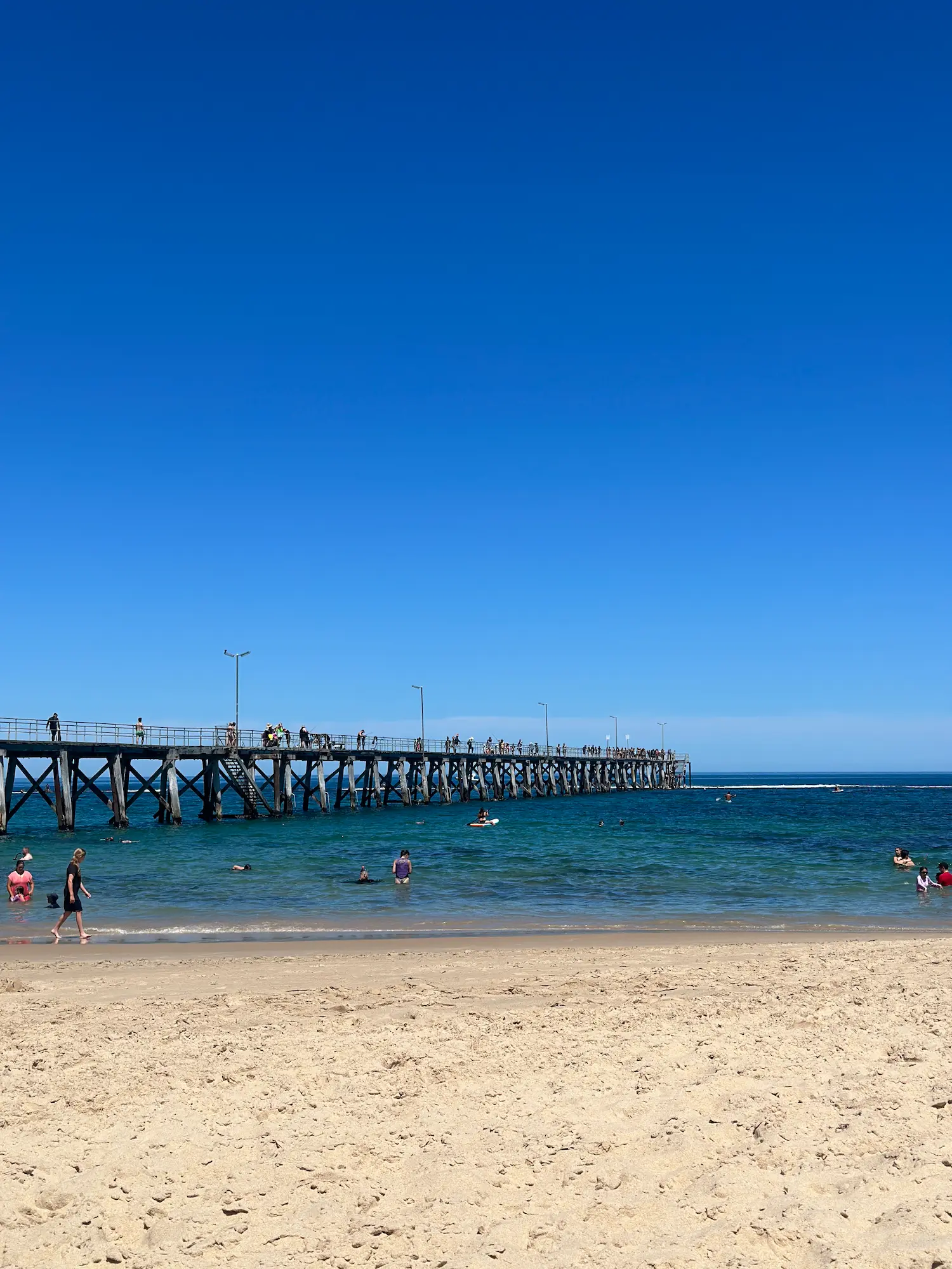 People swim and paddle in the clear turquoise water beside Port Noarlunga Jetty on a sunny day, with the long timber pier stretching out over the sea beneath a cloudless blue sky. Port Noarlunga is a popular coastal destination for Adelaide day trips, offering calm beaches, snorkeling, and relaxed seaside views.