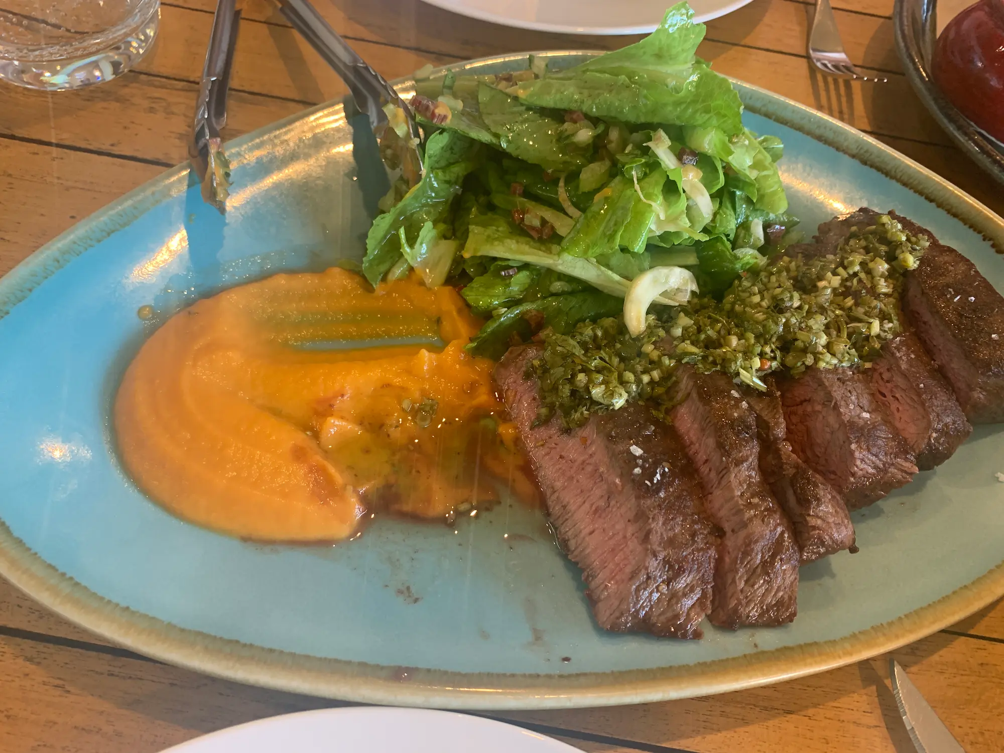 Sliced medium cooked steak topped with a fresh herb sauce is served with creamy pumpkin puree and a crisp green salad on a blue ceramic plate at Watervale Hotel. This beautifully presented meal showcases the regional dining experiences you can enjoy on scenic trips from Adelaide.