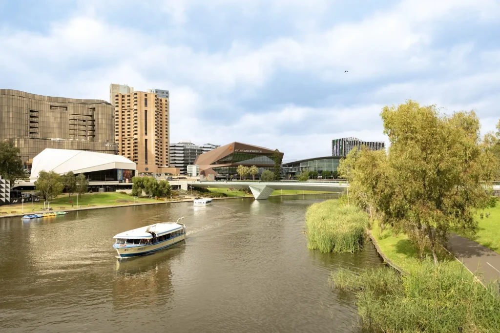 A sightseeing boat cruises along the River Torrens with the Adelaide Convention Centre, modern city skyline, and riverside parklands under a partly cloudy sky. A river cruise is one of the best things to do in Adelaide, offering a relaxing way to experience the city's waterfront and iconic landmarks.