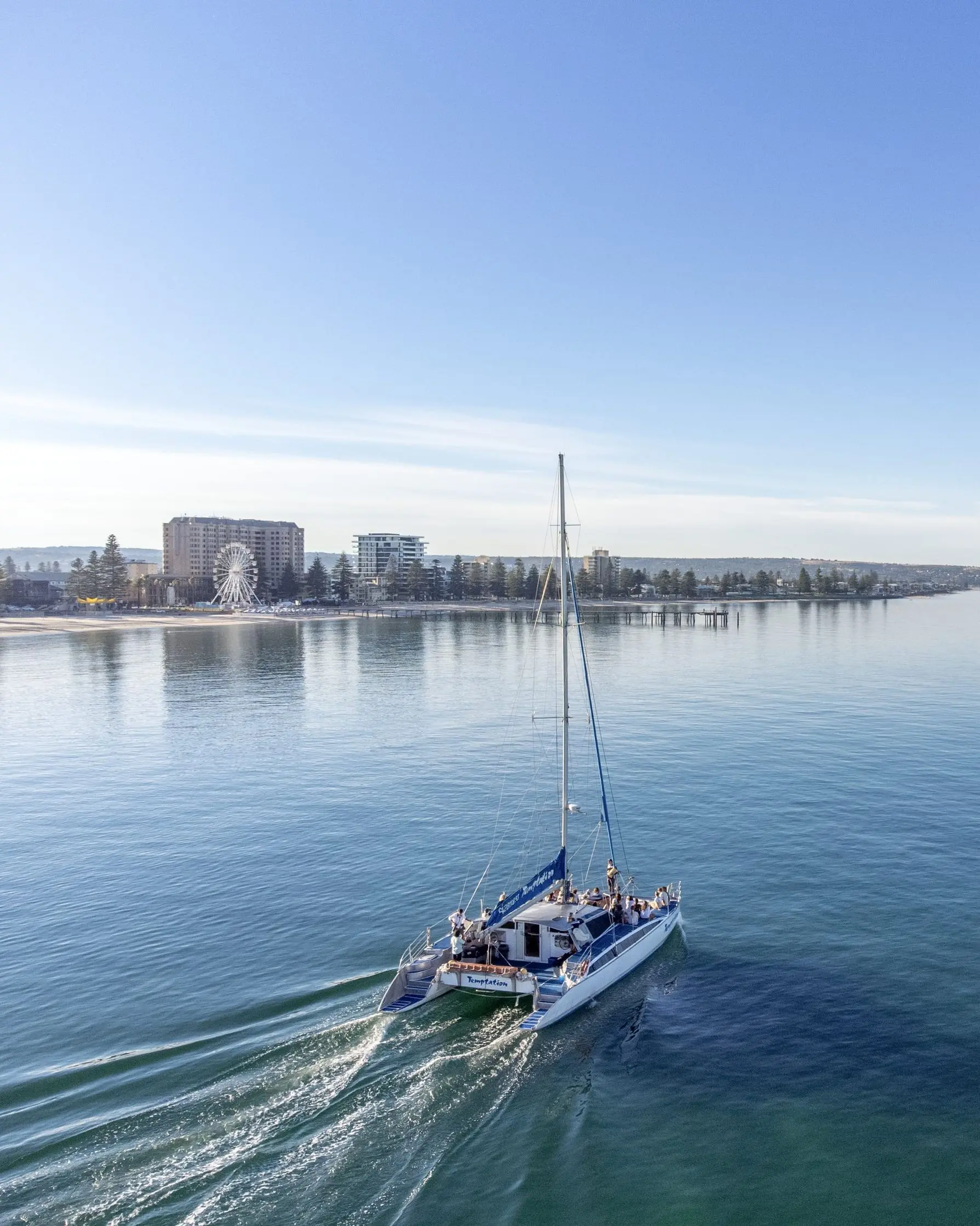 A catamaran sails across the calm waters of Glenelg with passengers on board while the shoreline, beach, and waterfront buildings stretch into the distance under a clear blue sky. A sunset cruise is one of the most relaxing Adelaide sightseeing experiences, offering beautiful coastal views from the water.