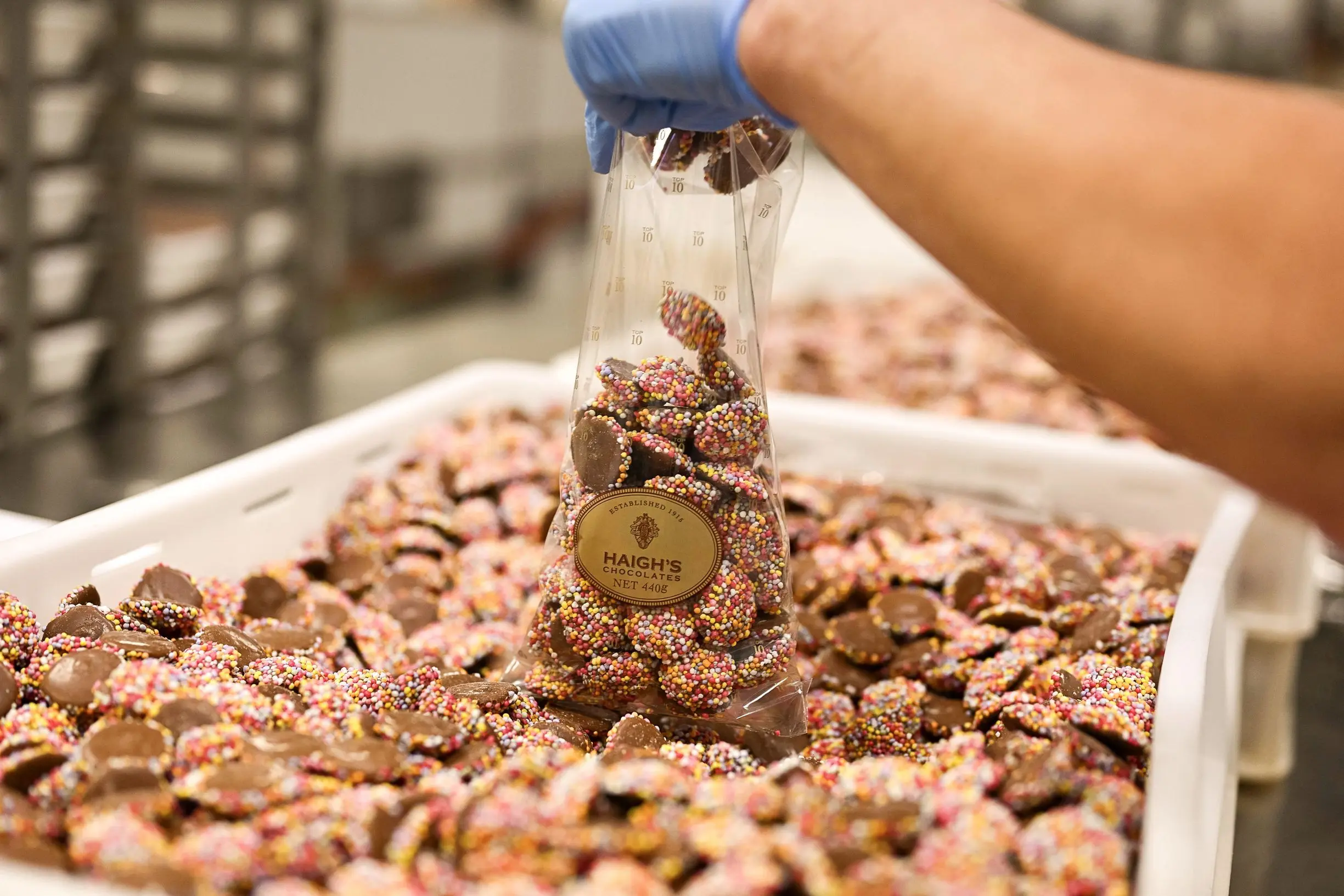 A staff member wearing a blue glove fills a bag with colorful chocolate freckles at the Haigh's Chocolates factory, with trays of freshly made chocolates in the background. A factory tour and chocolate tasting is one of the sweetest Adelaide experiences for visitors wanting to discover the city's famous confectionery.