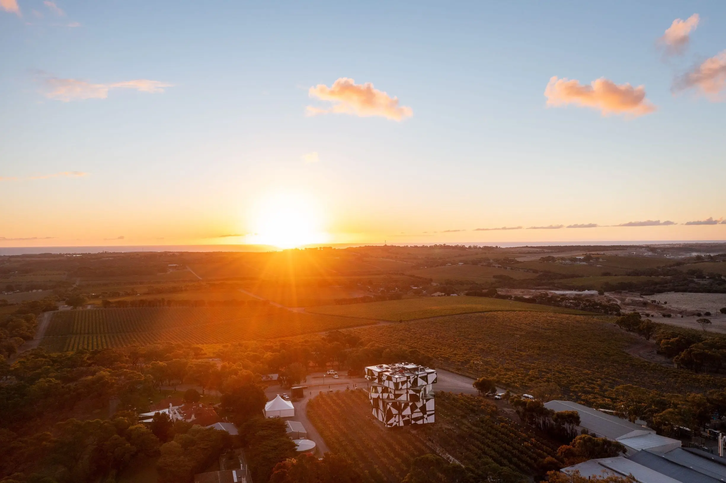 An aerial view captures the sun setting over rolling vineyards with the striking d'Arenberg Cube standing among the vines beneath a glowing orange sky. Visiting this iconic winery is one of the best answers to what to do in Adelaide for spectacular views, wine tasting, and unique architecture.