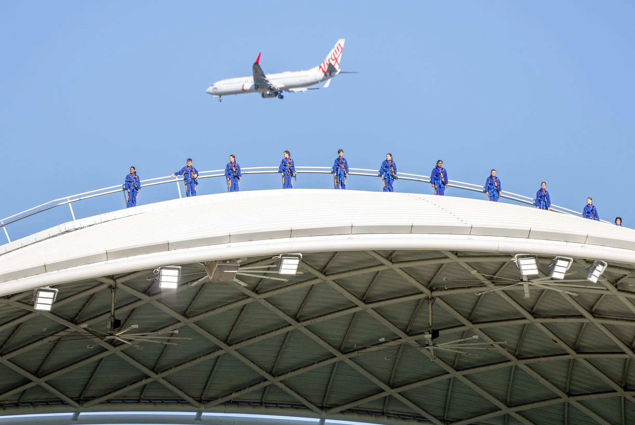 A group of people in blue jumpsuits walks across the curved roof of Adelaide Oval while a Virgin Australia plane flies overhead against a clear blue sky. RoofClimb Adelaide is one of the most exciting Adelaide activities for panoramic city views and a unique perspective of the iconic stadium.
