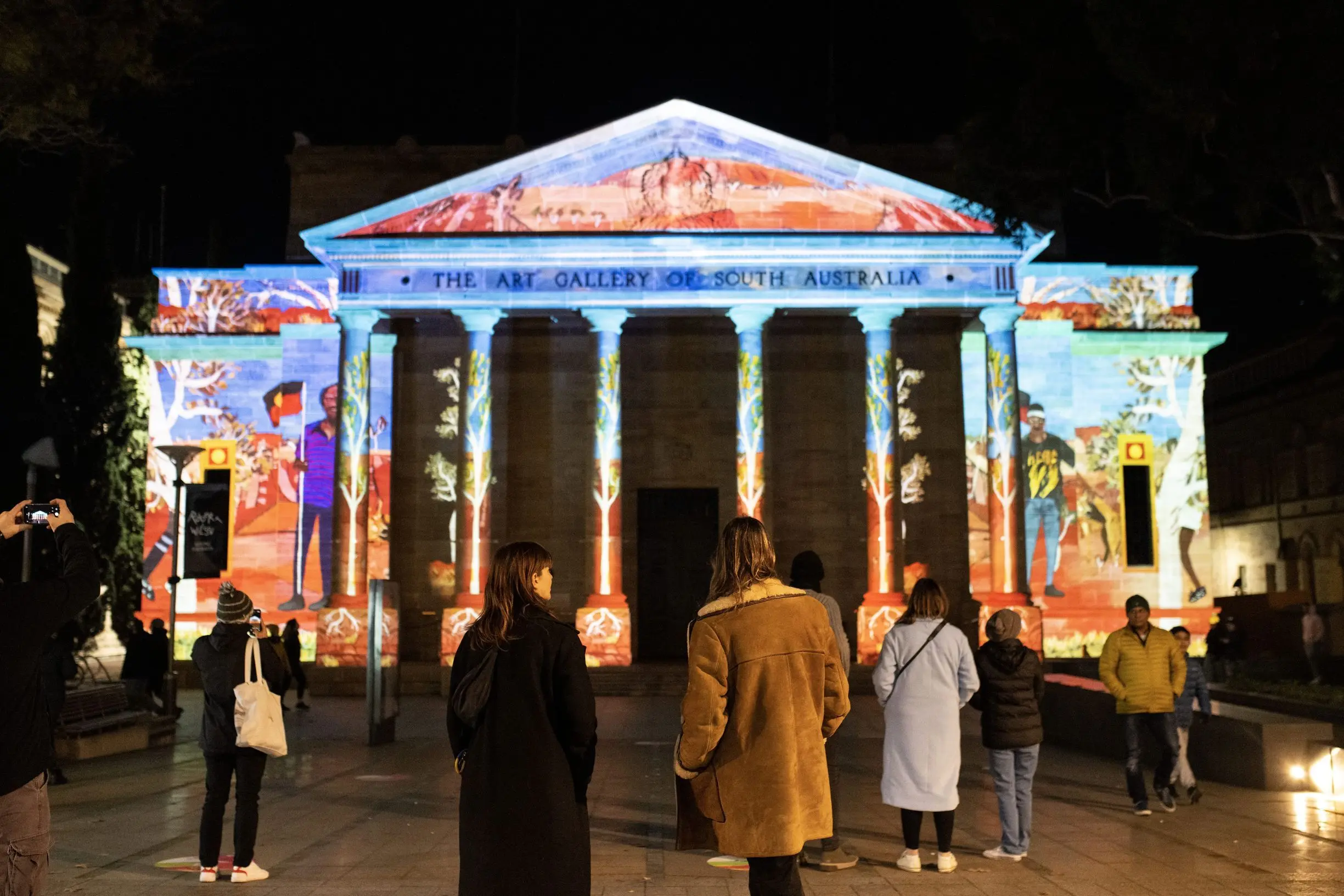 Visitors stand in front of the Art Gallery of South Australia at night as vibrant projection art transforms the building's historic facade with colorful illustrations and landscapes. This large scale light display is one of the most memorable Adelaide attractions for experiencing the city's arts and culture after dark.