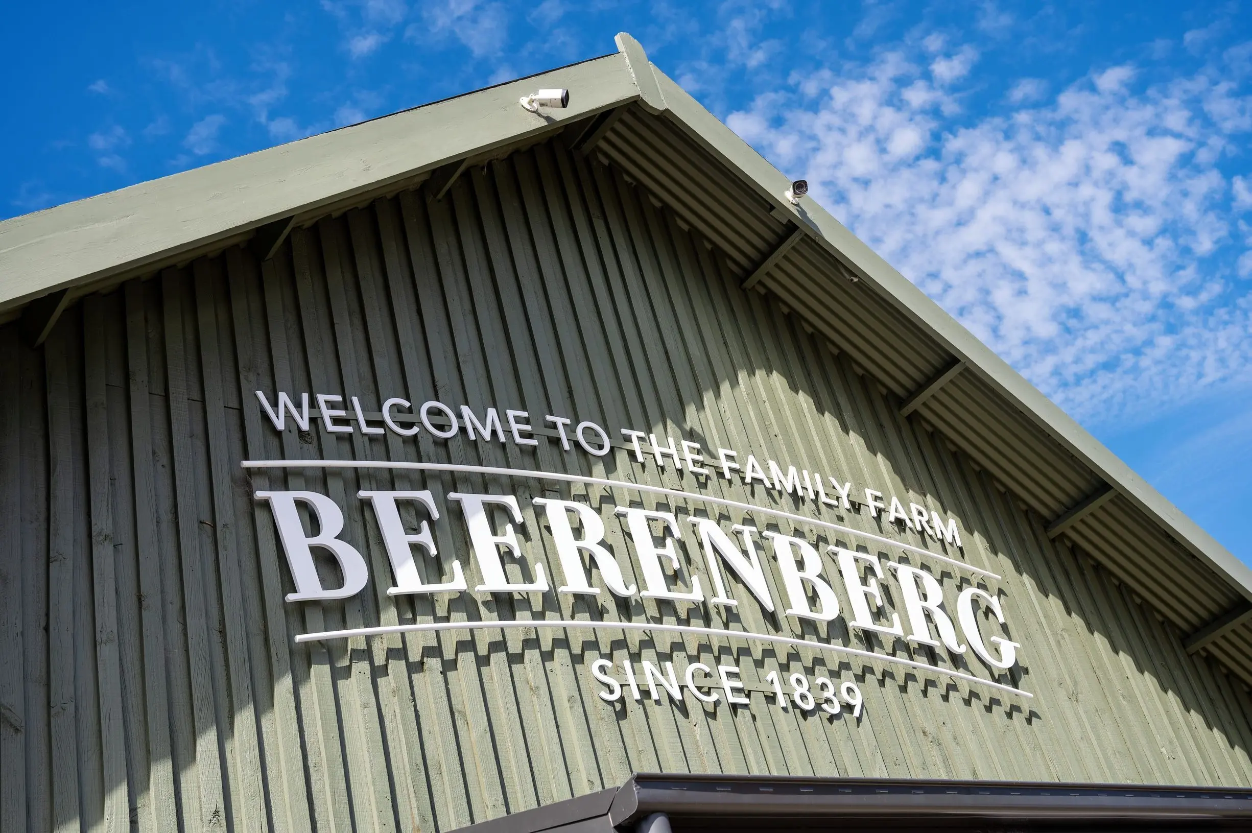 The entrance to Beerenberg Farm features large white lettering reading "WELCOME TO THE FAMILY FARM BEERENBERG SINCE 1839" on a green barn style building beneath a bright blue sky. Visiting Beerenberg Farm is one of the must see places in Adelaide for picking fresh strawberries and tasting locally made jams and produce.