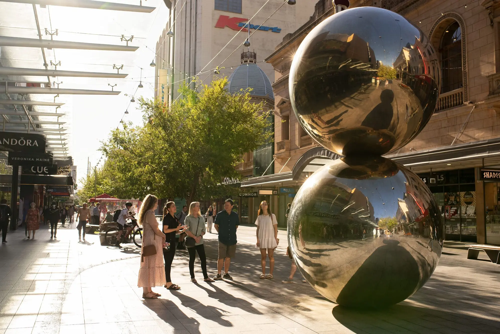 Shoppers gather around the large mirrored Spheres sculpture in Rundle Mall as afternoon sunlight reflects across the polished surface. The pedestrian shopping street offers a glimpse of everyday city life for visitors exploring Adelaide.