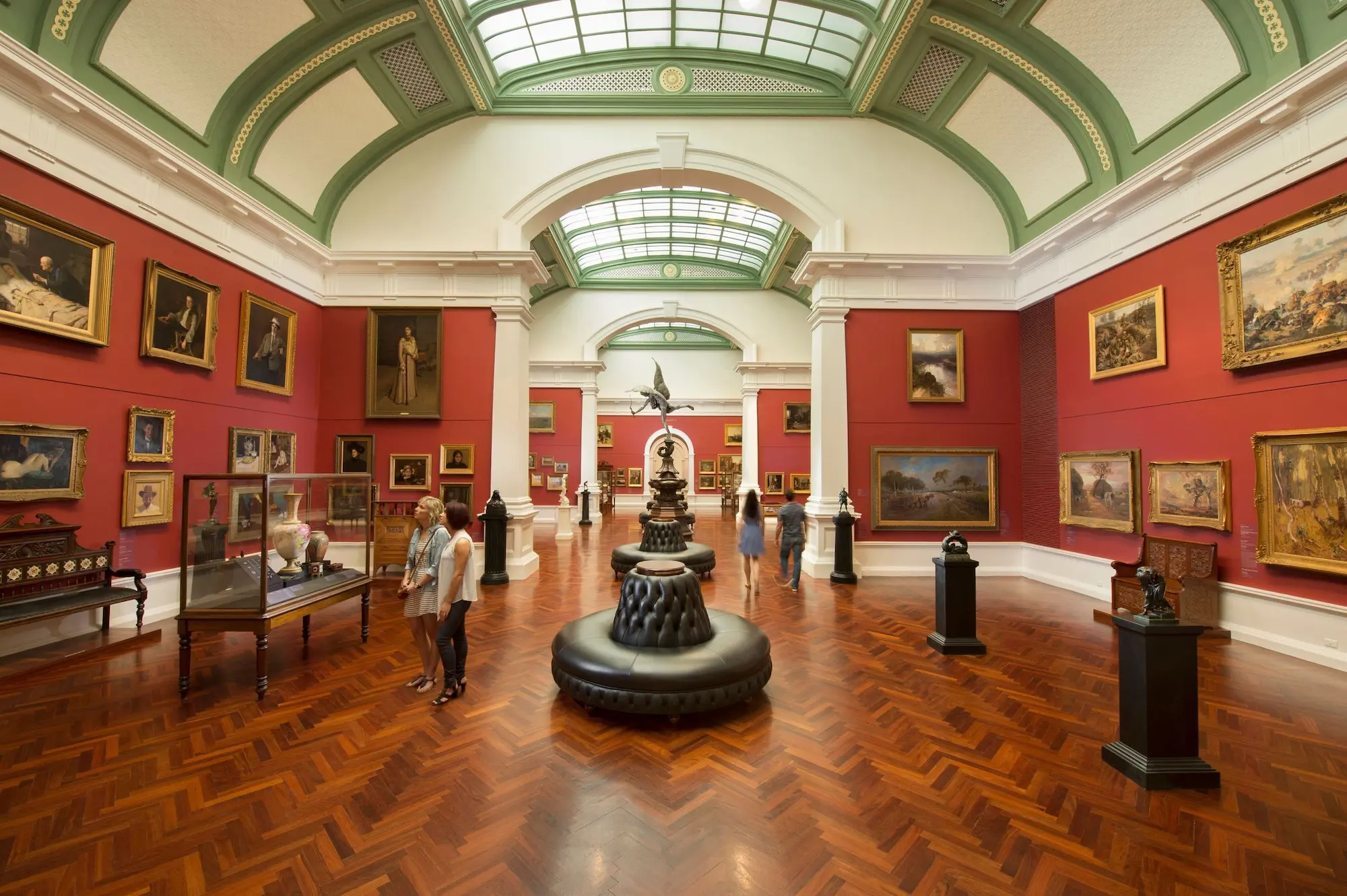 Grand interior of the Art Gallery of South Australia with red walls covered in paintings. polished parquet floors. arched ceilings. and visitors browsing the collection. The free gallery is one of the cultural attractions to discover during 3 days in Adelaide.
